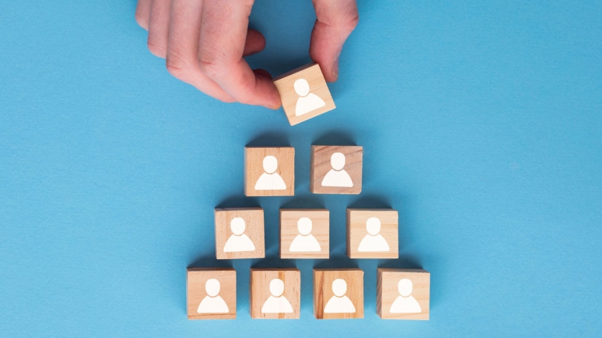 A hand places a wooden block with a person icon on top of a pyramid made from similar blocks, all on a blue background, symbolizing hierarchy or organizational structure.