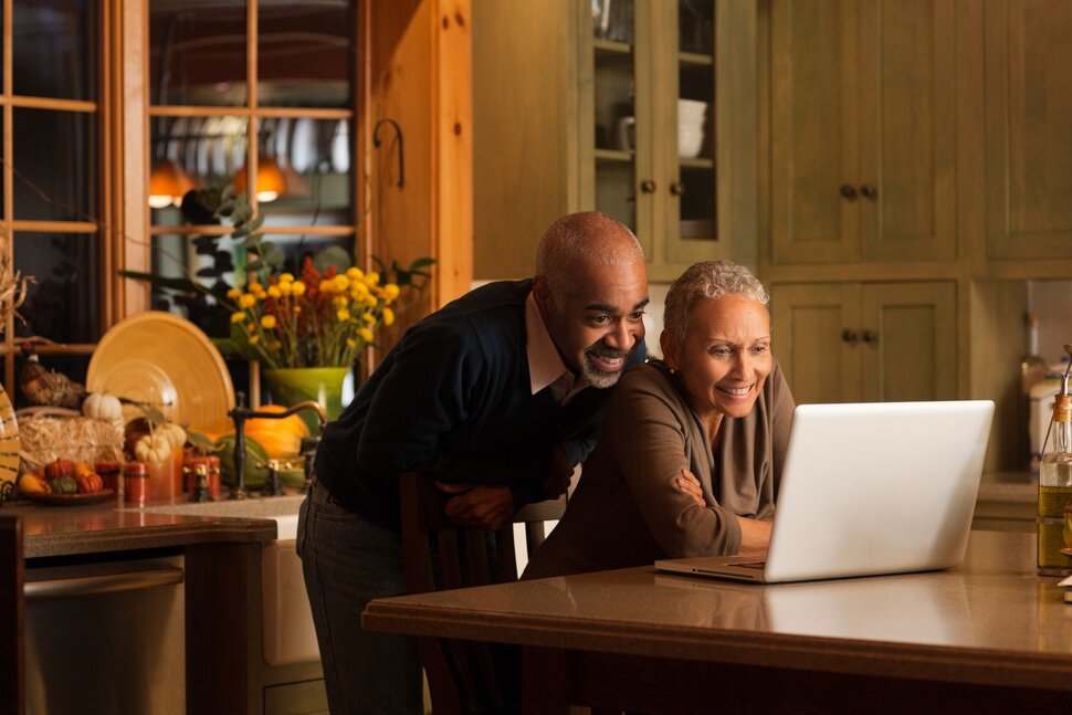 An older couple smiles and looks at a laptop together in a warmly lit kitchen, surrounded by dishes, flowers, and wooden cabinets.